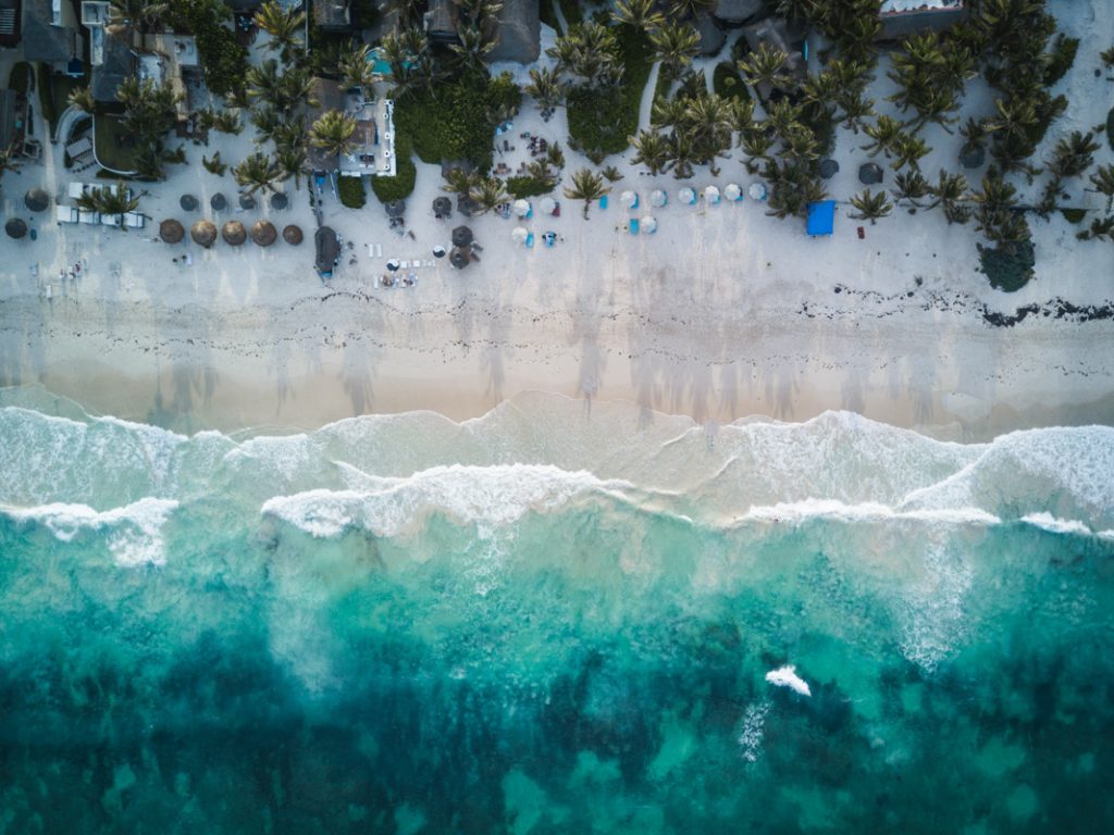 Drone shot of a breathtaking beach in Tulum, Mexico, featuring powdery white sand, crystal-clear turquoise waters, and lush green palm trees, providing a mesmerizing tropical paradise