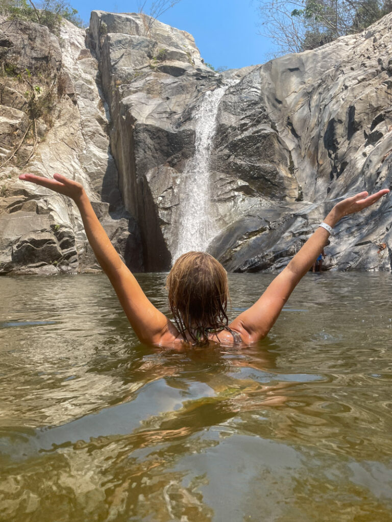 girl swimming holding her hands up in front of quimixto waterfall