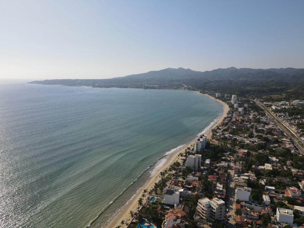 aerial shot of main beach in bucerias from above. the calm ocean meets the sand with the town behind it.