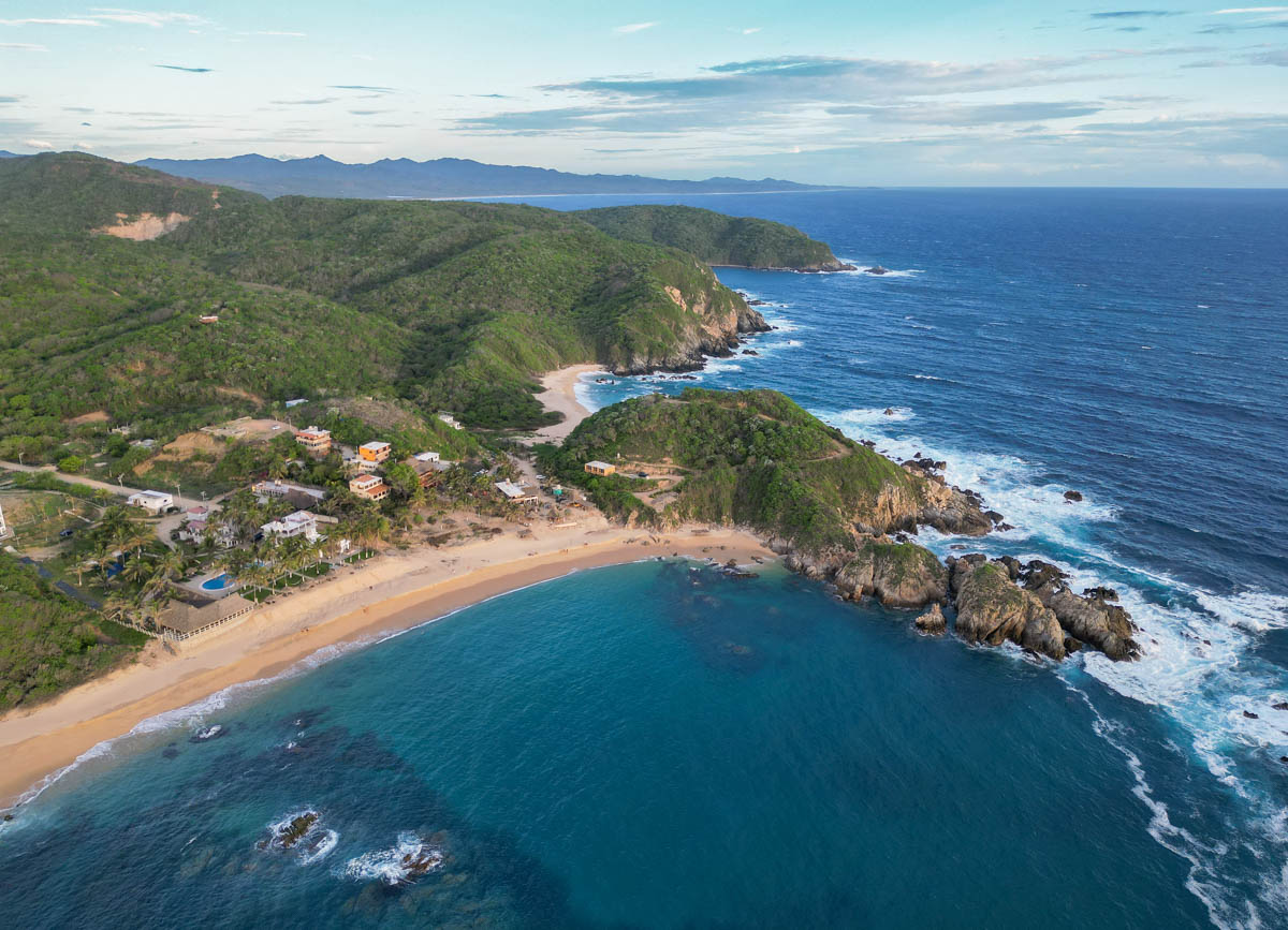 Aerial view of a coastal landscape near Puerto Vallarta, with sandy beaches, rocky shoreline ideal for snorkeling, turquoise water, and green hills under a partly cloudy sky.
