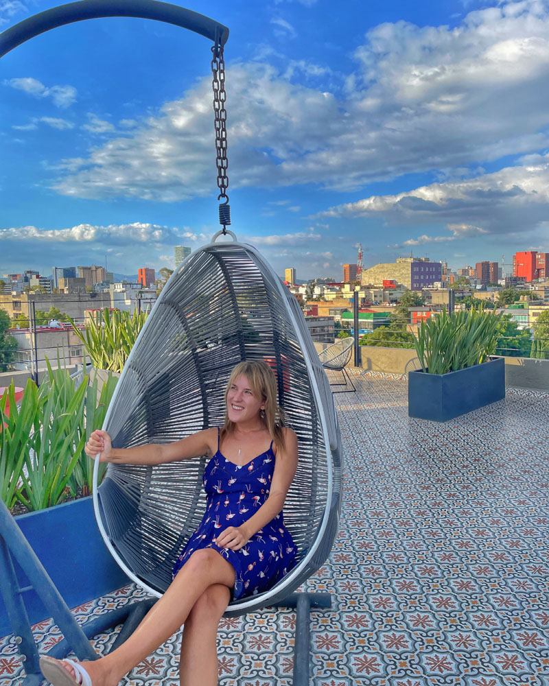 lora sitting on a chair smiling on a rooftop in mexico city. you can see the city skyline in the background.