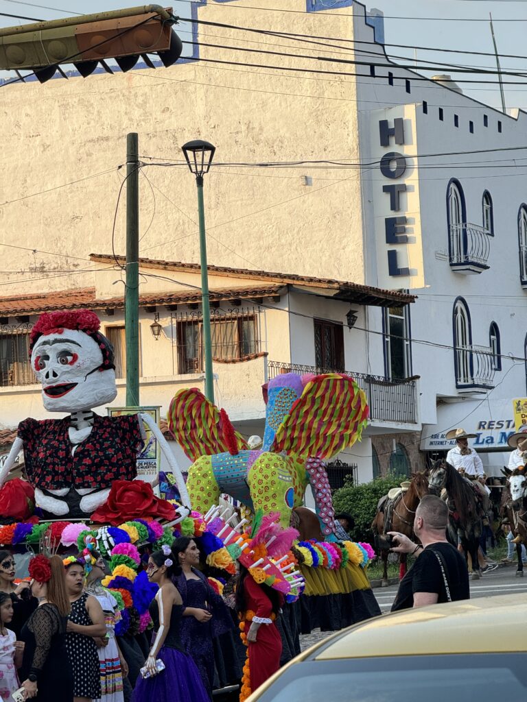 A festive Day of the Dead parade with colorful costumes and a large skeleton display moves through a street in Puerto Vallarta, lined with buildings under a "Hotel" sign.