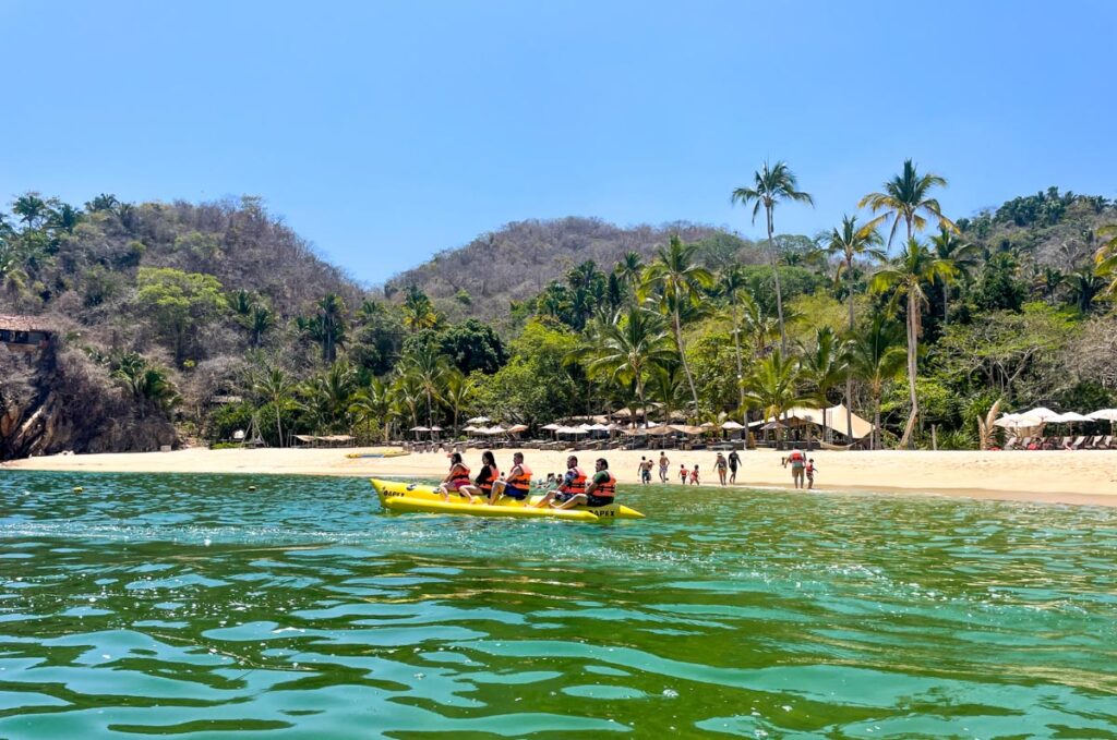 five tourists on a banana boat in turquoise water in puerto vallarta mexico. behind them is a white sand beach and mountains.