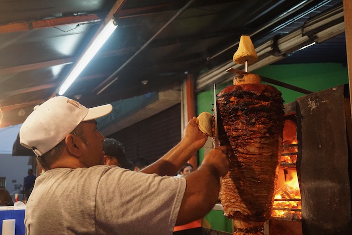 A skilled man meticulously scrapes tender beef onto a rotating spit, preparing the flavorful meat for authentic tacos al pastor. 