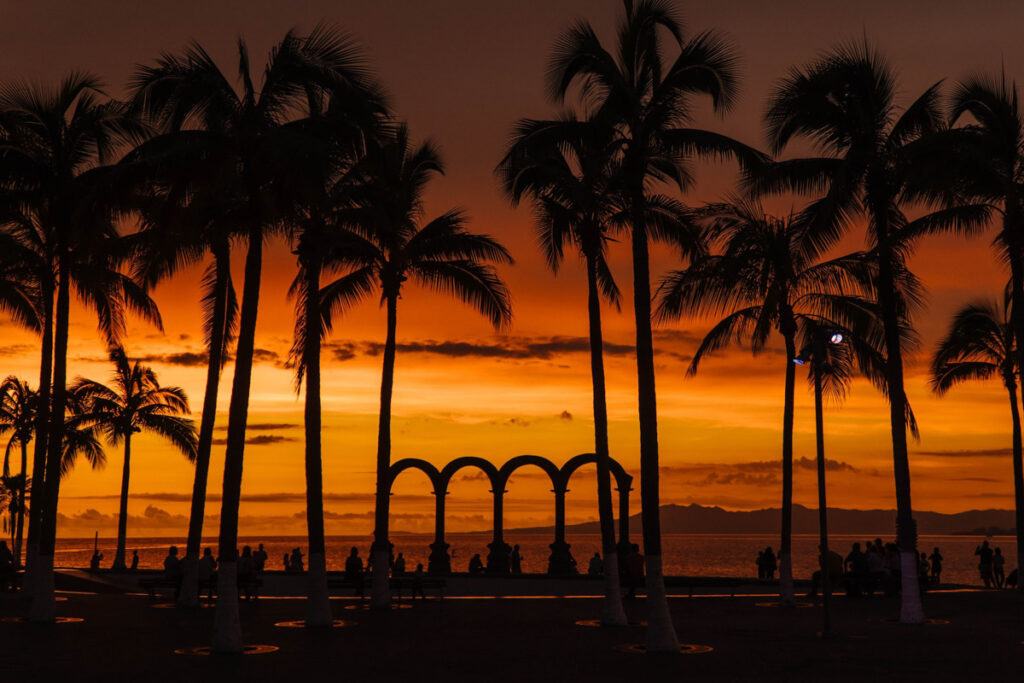 a beautiful sunset with yellow, orange and red tones on the malecon. silhouettes of people standing by palm trees facing the ocean.