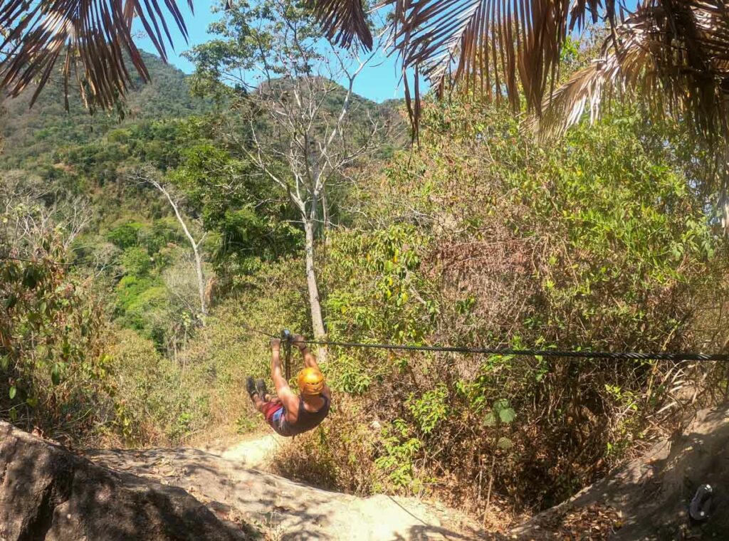 An adventurous man exhilaratingly ziplines through the lush green jungle of Puerto Vallarta, surrounded by towering trees and a breathtaking natural landscape.