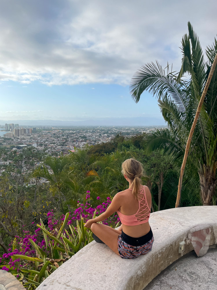 lora sitting at an outdoor yoga studio with purple flowers and palm trees around her, looking towards the city of puerto vallarta