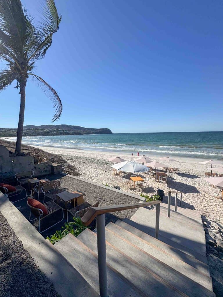 Concrete steps lead down to a sandy beach with tables, chairs, and umbrellas set up. A palm tree stands on the left, and the ocean and hills—reminiscent of towns near Puerto Vallarta—are visible in the background.