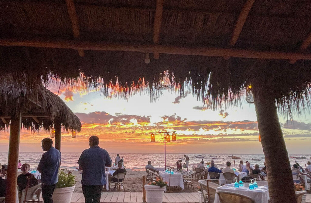 The palapa at El Dorado offers a picturesque setting with its thatched roof and panoramic view. As the sun sets on the horizon, casting a soft pink hue across the sky, guests seated beneath the palapa savor their meals. 