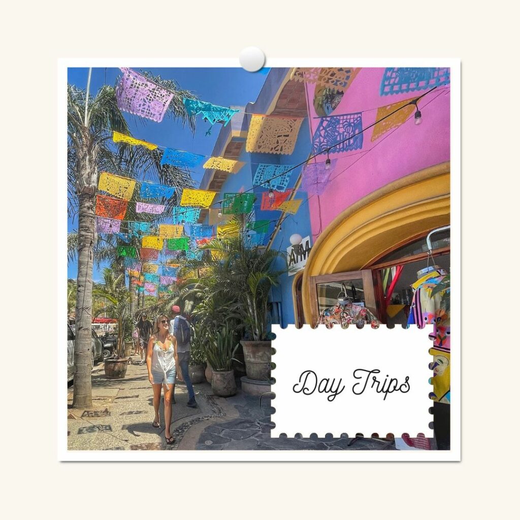 lora is walking down a colorful street in sayulita mexico. the flags are yellow blue and purple next to a pink and blue building. she is smiling.