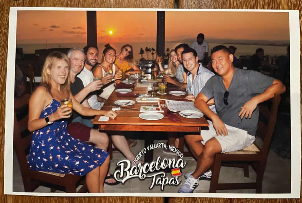 A jovial group of friends gathers at Barcelona Tapas, a vibrant restaurant renowned for its delectable Spanish cuisine. Against the backdrop of a radiant orange sunset, they revel in the lively atmosphere, sharing tapas and raising their glasses in celebration