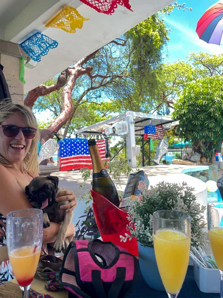 lora holding a pug at casa capula brunch. behin her is a drag performer and in front of her is a mimosa and bottle of champagne