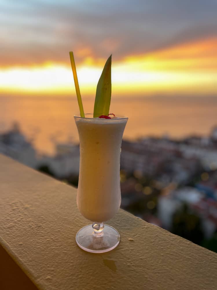 a pina colada cocktail in puerto vallarta sitting on a ledge overlooking the city at sunset