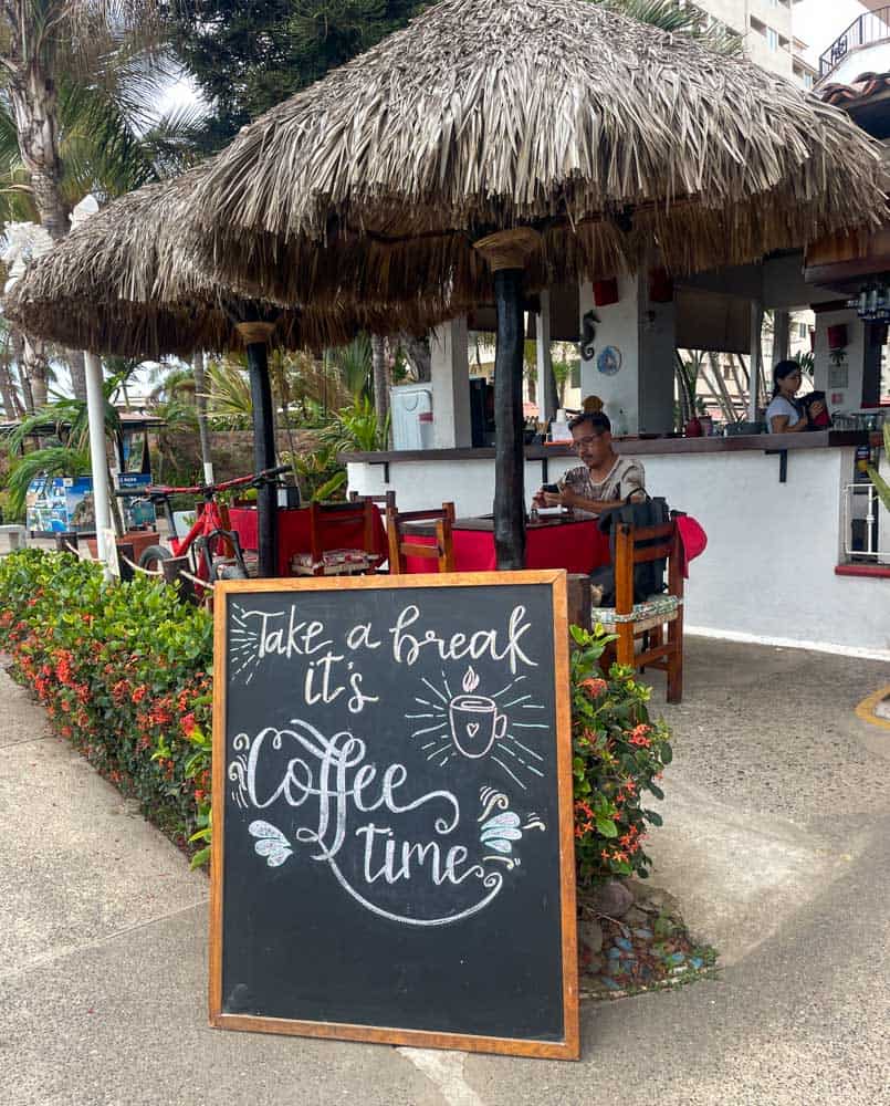 take a break and enjoy some coffee time at a coffee shop along the Malecon in Puerto Vallarta, as indicated by a sign, while overlooking the picturesque view.