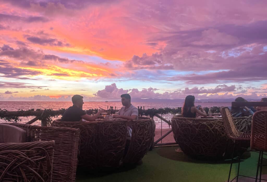 A group of people is seen dining at Ik Restaurant, a chic establishment situated along the coast of Puerto Vallarta. As the sun sets, casting a mesmerizing purple hue across the sky, patrons delight in their meals, their faces illuminated by the warm glow, creating a captivating scene of culinary bliss and natural beauty