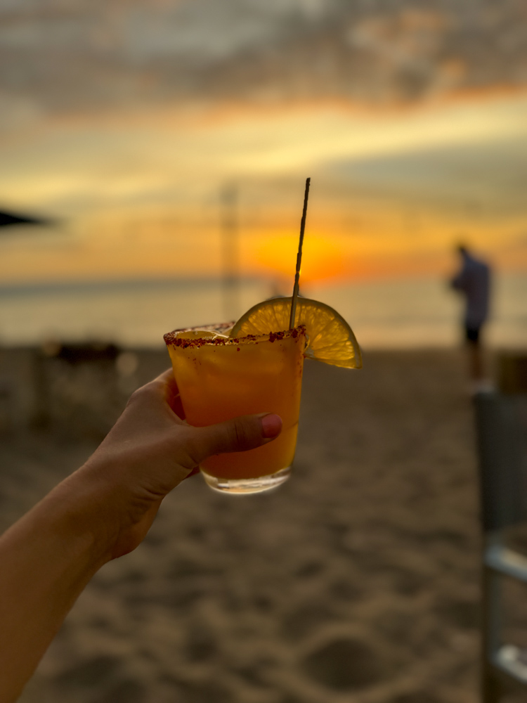 hand holding cocktail in front of beach with blurred sunset in backdrop