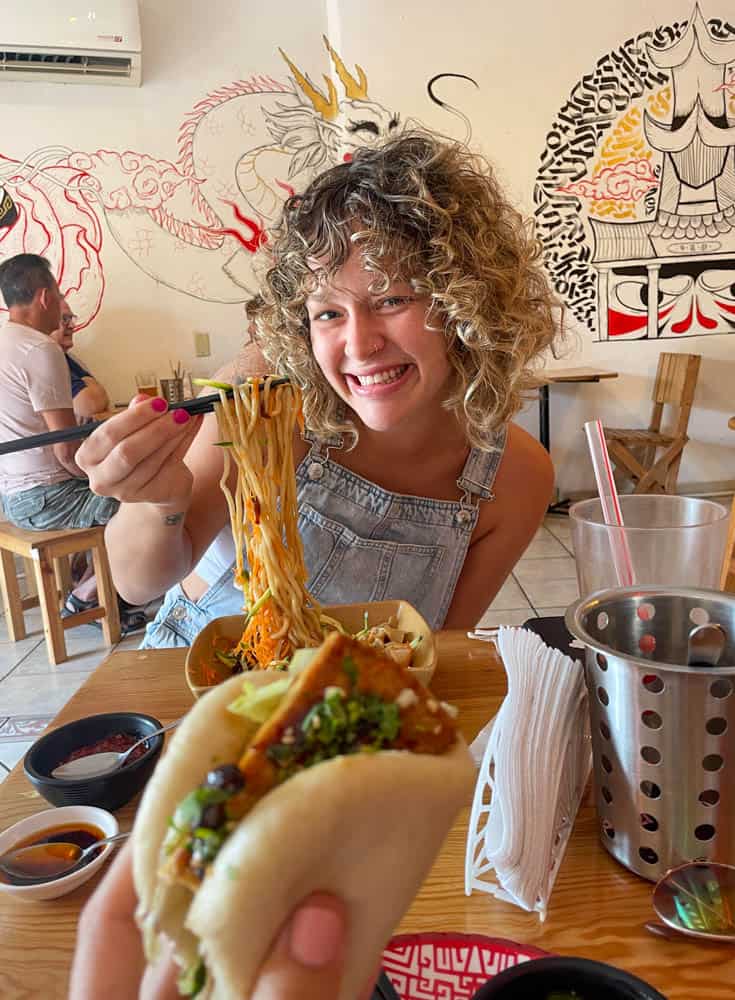 a hand holding a bao sandwhich wiht a women in the background smiling while holding noodles she is about to eat