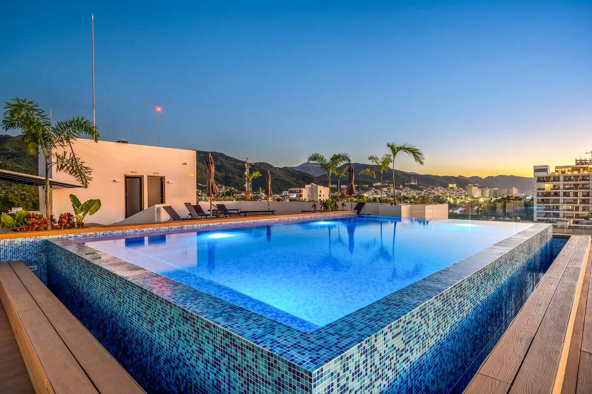 Rooftop infinity pool with mosaic tiles, surrounded by wooden decking, lounge chairs, palm trees, and cityscape view at sunset.